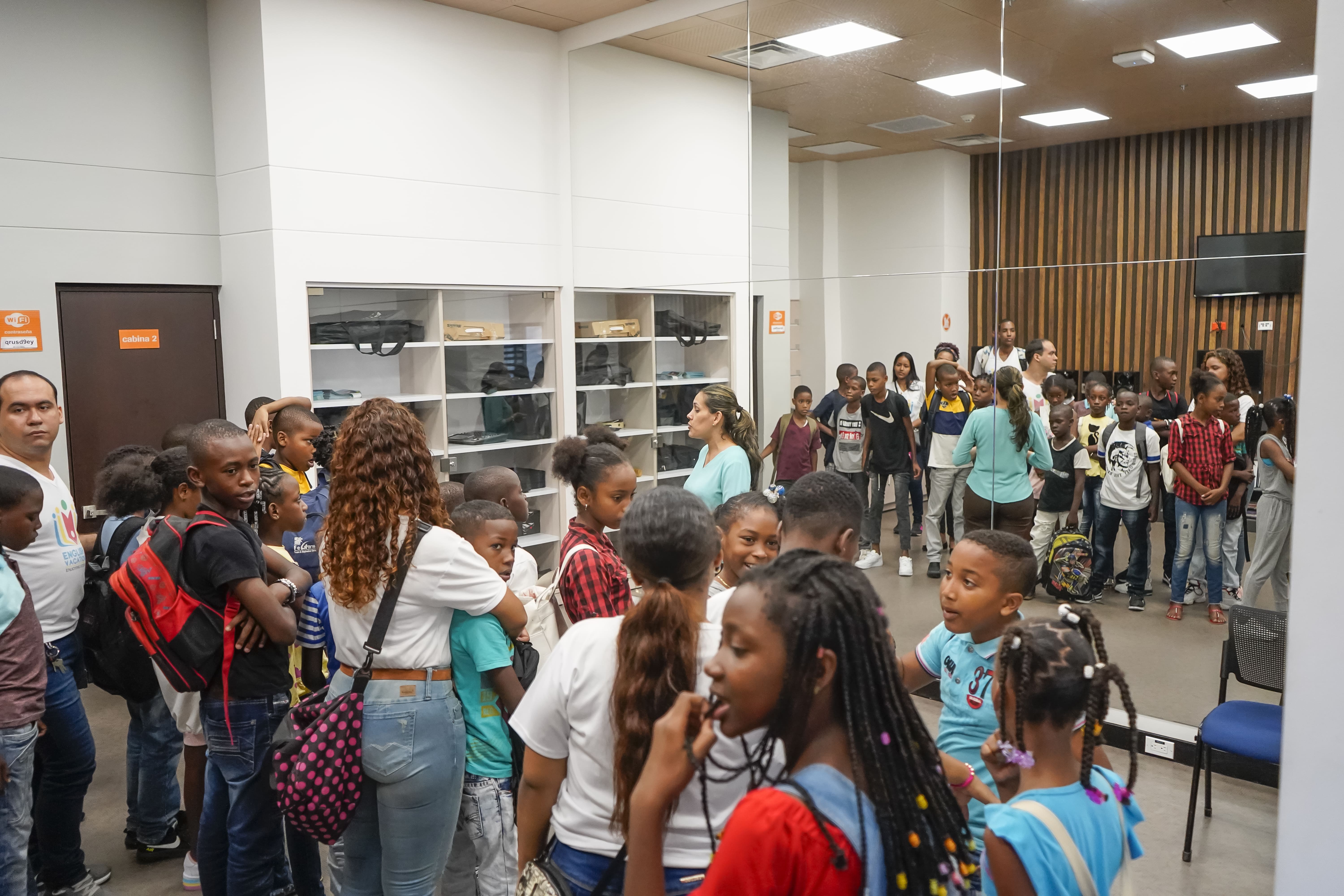 La imagen puede contener a niños,jovenes y adultos de pie, reunidos escuchando una charla dirijida por una mujer que se encuentra alfrente de ellos.